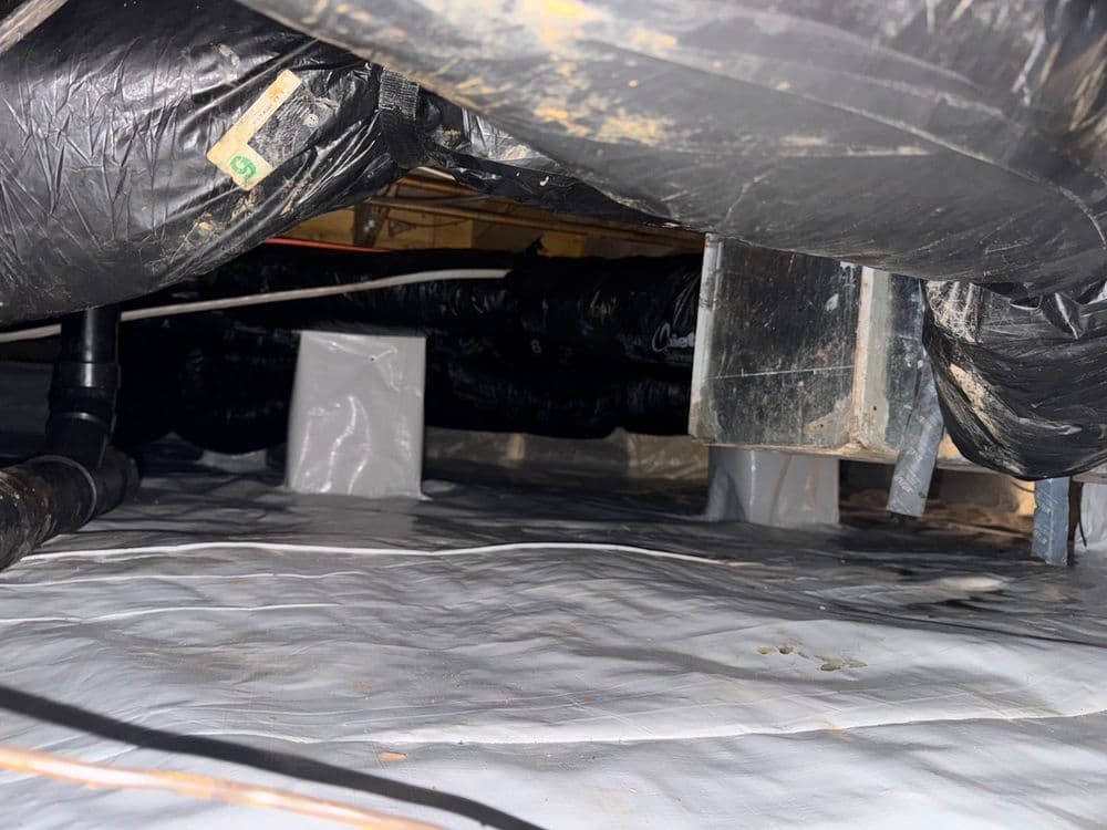 Crawl space view with black plastic ducts and white moisture barrier on the ground.