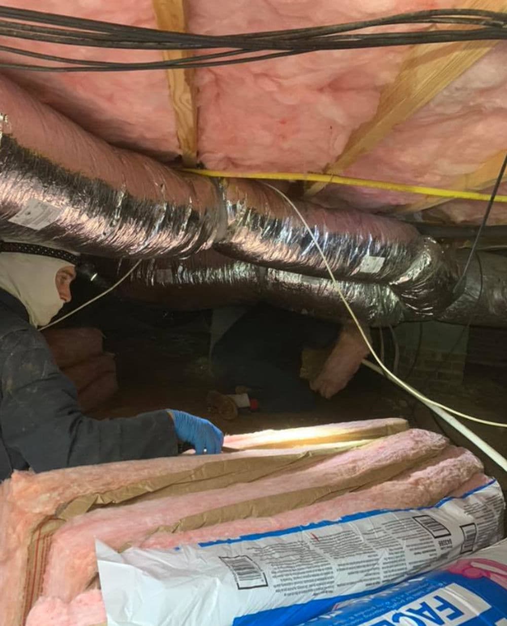 Worker inspecting ductwork in an attic with pink insulation and cardboard materials.