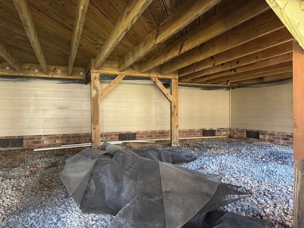 Interior view of a wooden structure with exposed beams, gravel floor, and construction materials.