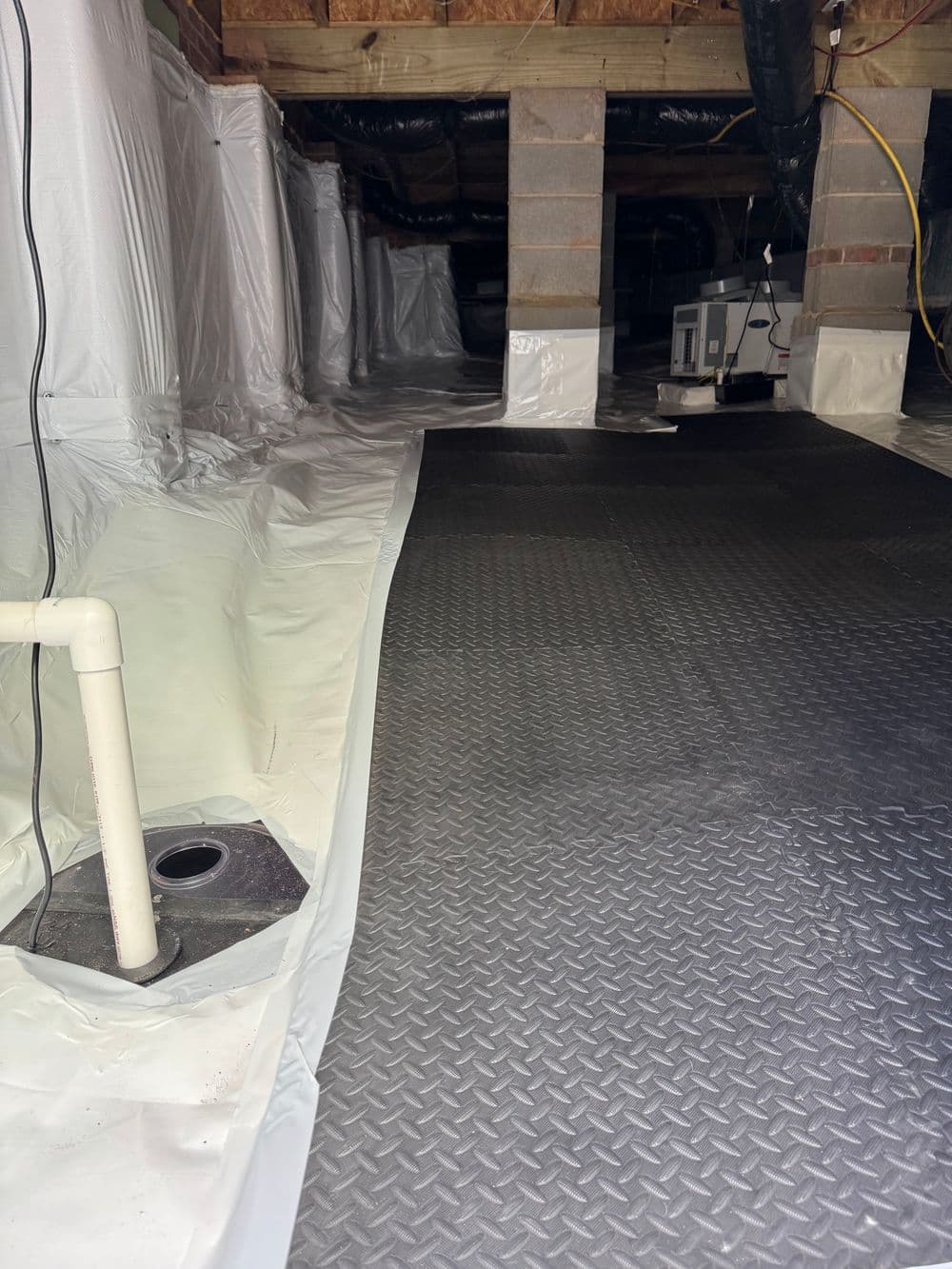 Crawl space with white plastic vapor barrier and gray drainage flooring for moisture control.